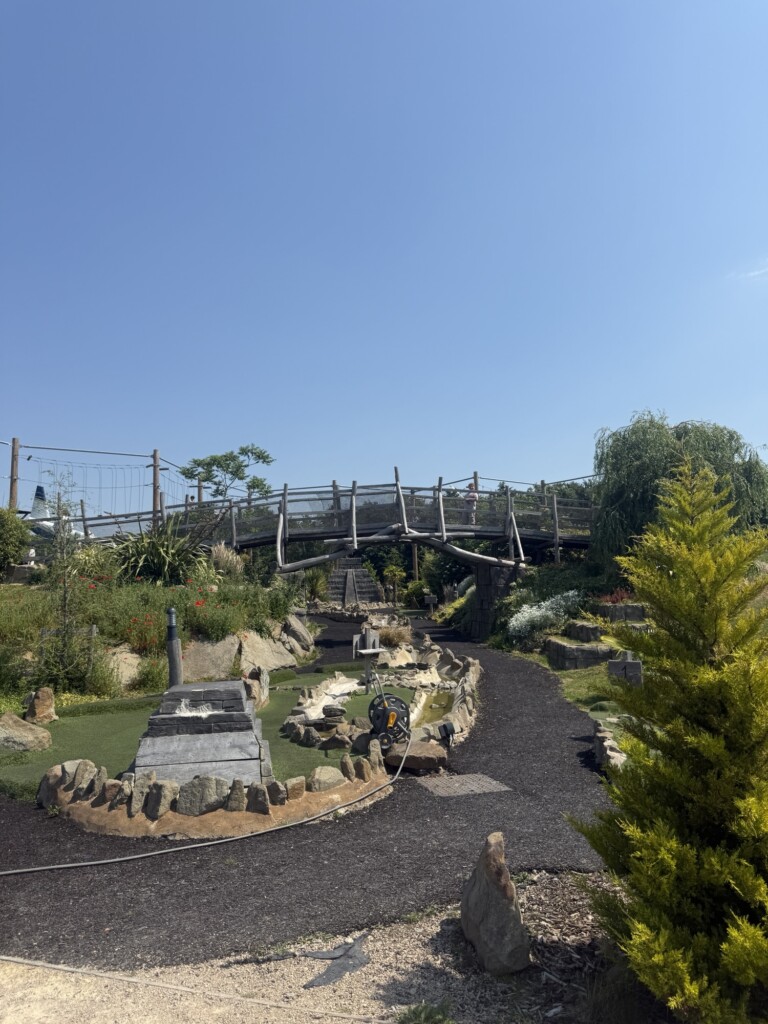 A peaceful garden with curving paths, rocks, a pond, and a wooden bridge beneath a clear blue sky.