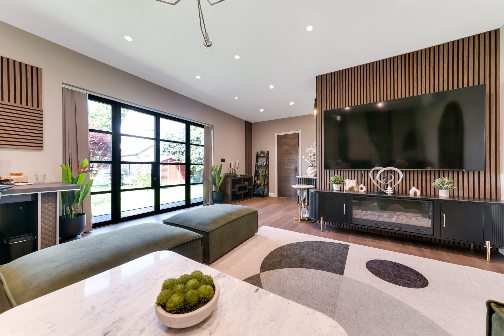 A sitting room with a large telly, black cupboards, a geometric rug, plants, and tall windows looking out to a garden.
