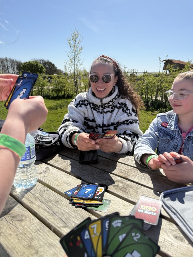 Three people play UNO No Mercy at a picnic table outside on a sunny day, with one smiling and the game box visible.