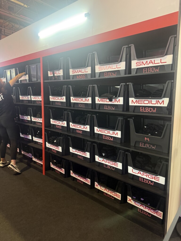 Black bins labelled for different sizes and types of protective gear are on shelves, with a person partly visible to the left.