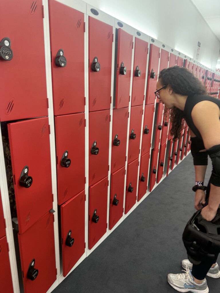 Someone with a helmet stands in a locker room, facing red lockers, one of which is slightly ajar.