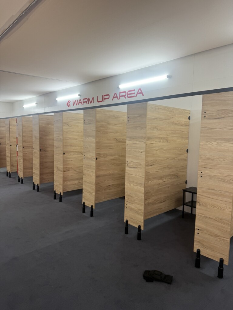 Several wooden changing cubicles with stools sit under a "WARM UP AREA" sign, and a lone sock rests on the carpeted floor.