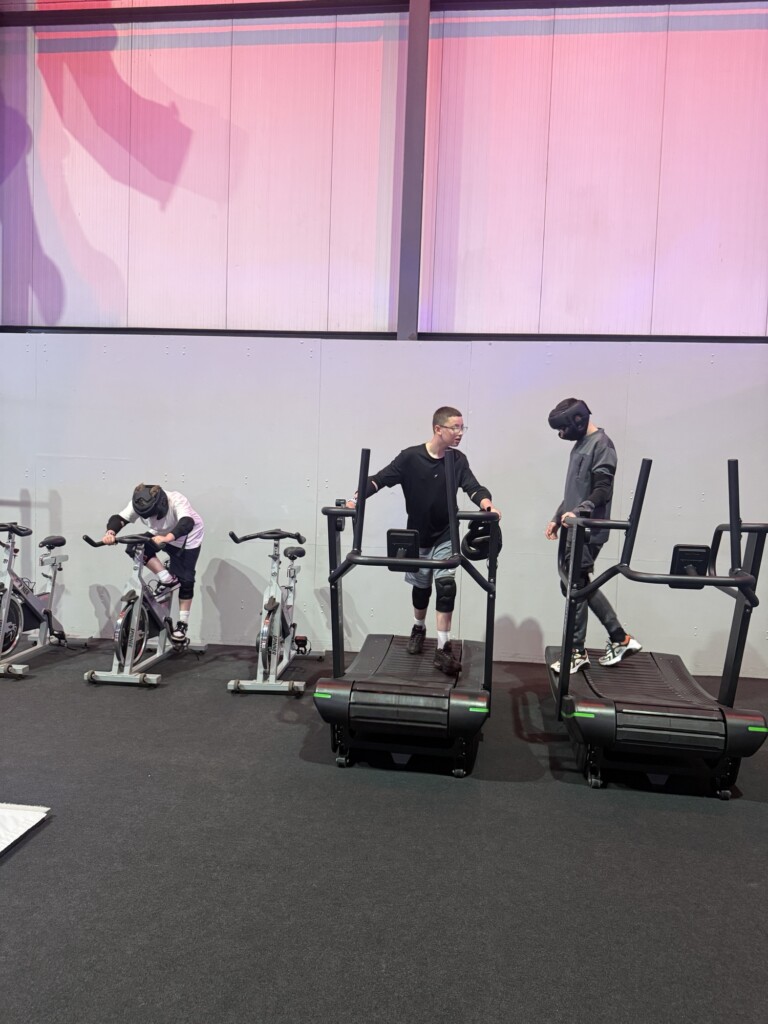 Three helmeted people exercise, two on bikes and one on a running machine, while another person stands nearby.