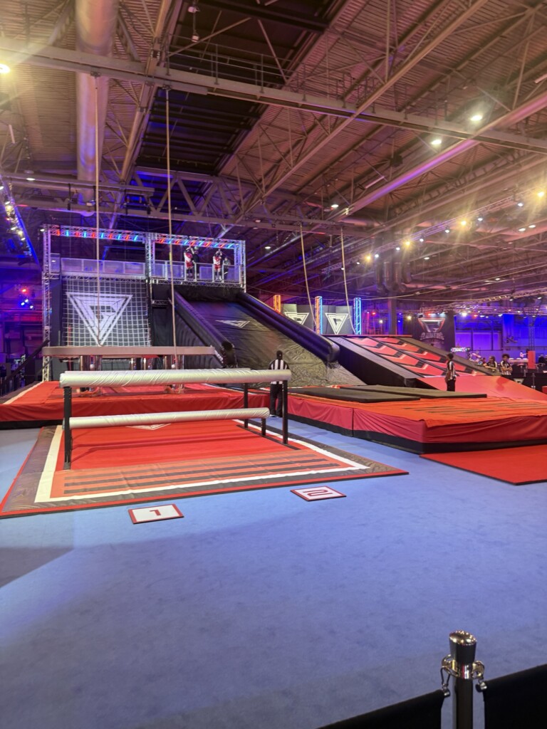 A gym with ramps, padded obstacles, and climbing frames on red mats and blue floors under bright indoor lights.