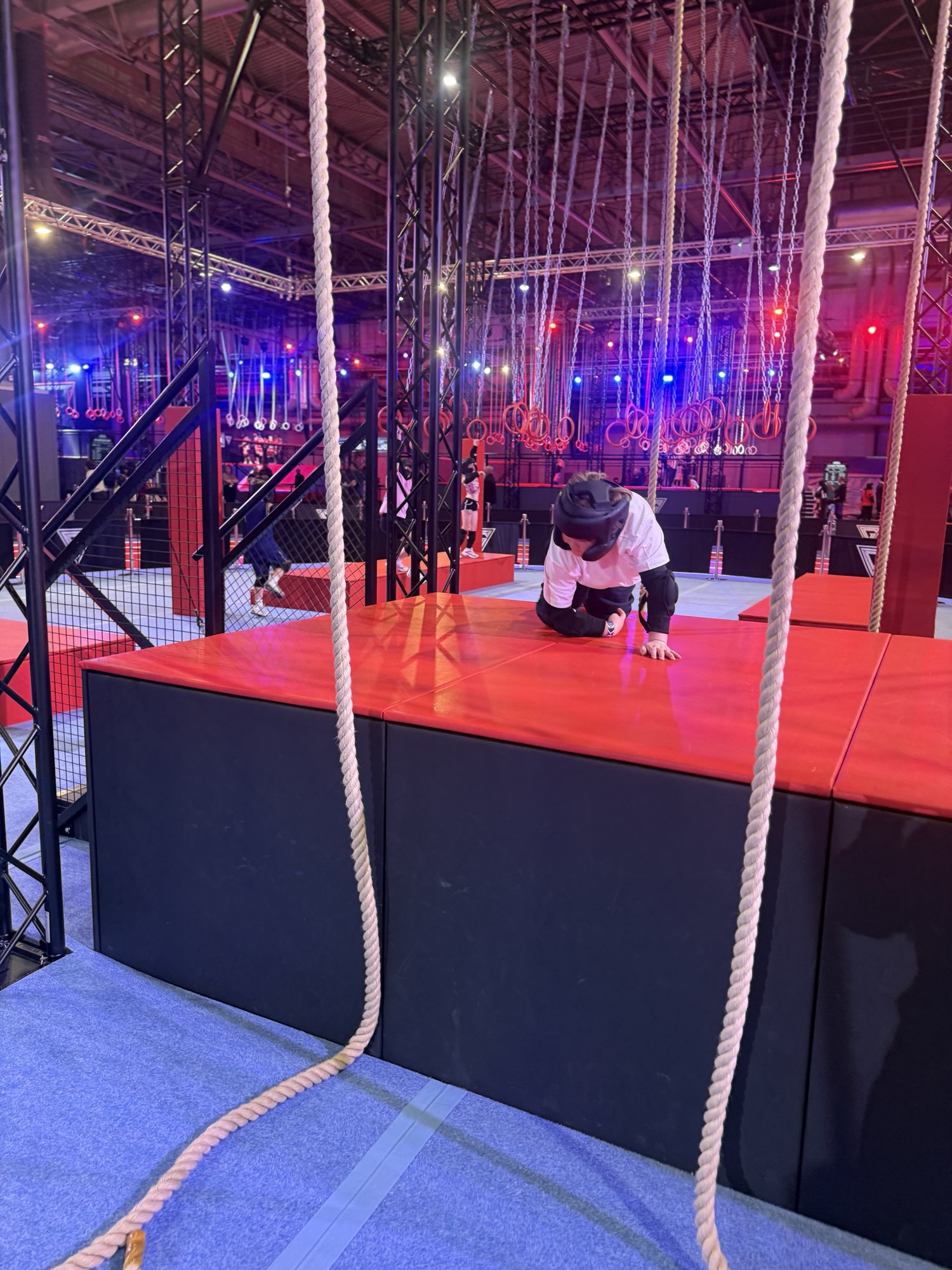 Someone wearing sports clothes and a helmet crawls on a red mat in an indoor obstacle course with ropes and metal frames.
