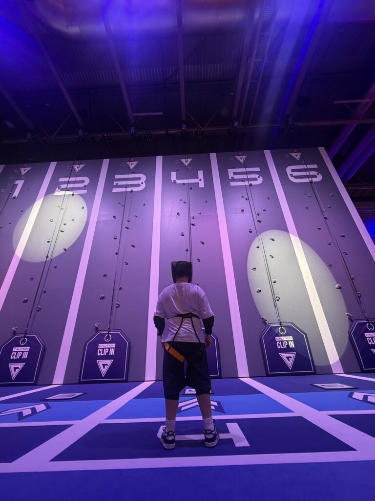 Someone in a harness stands in front of a numbered climbing wall, illuminated by purple light.