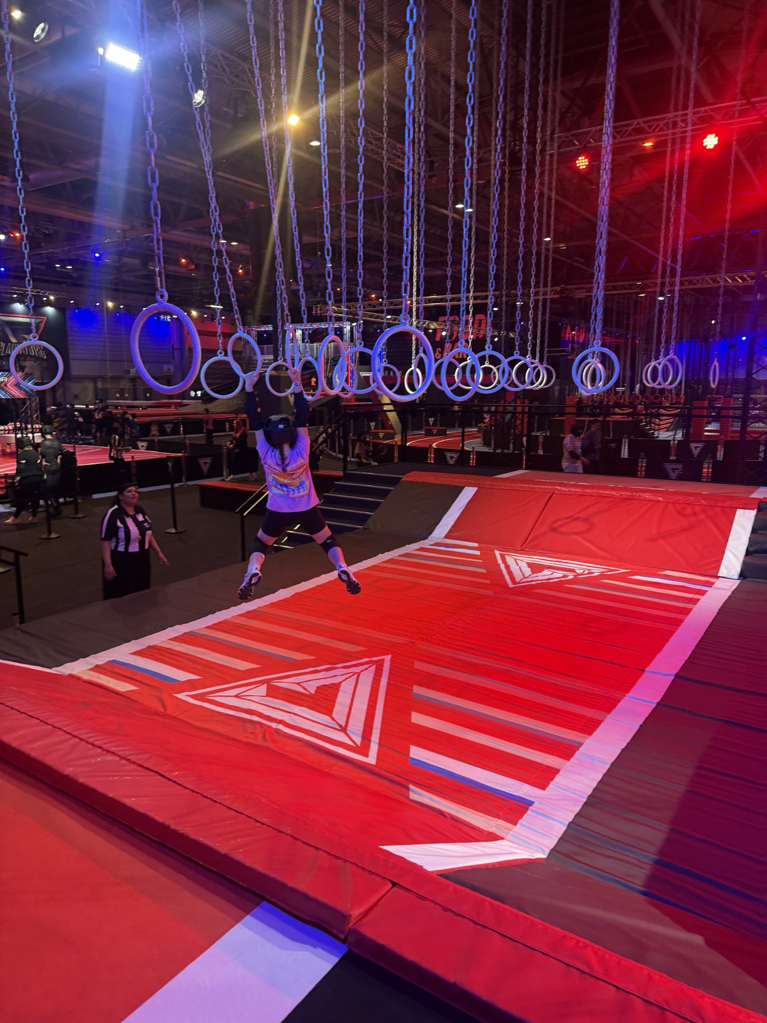 A child swings on rings above a red padded course as a referee watches in a brightly lit indoor arena.