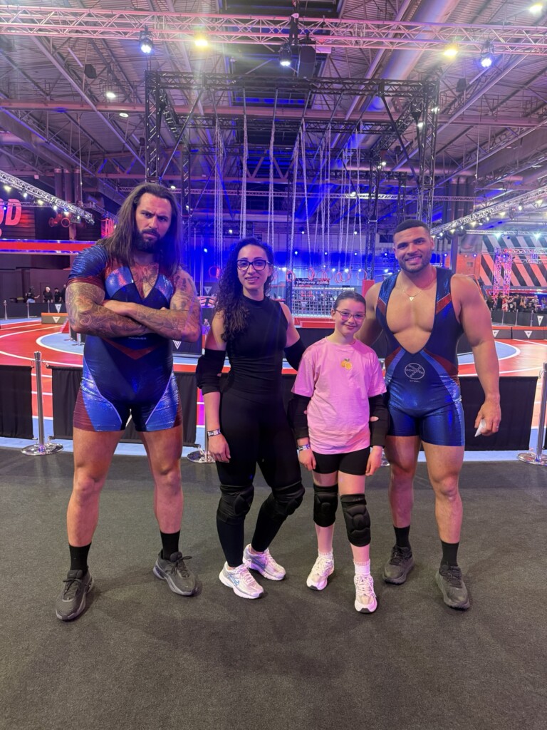Two men in blue and two women, one in black and one in pink, stand together on an arena floor under bright stage lights.