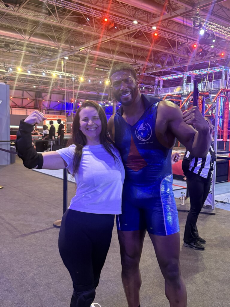 A smiling woman and man flex their arms together in a brightly lit indoor sports arena.