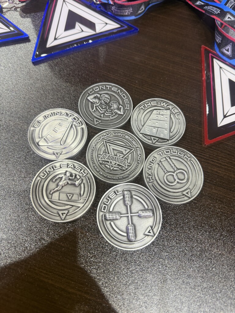Six silver medals with different event names lie on a dark wooden surface, each with a ribbon lanyard placed above them.