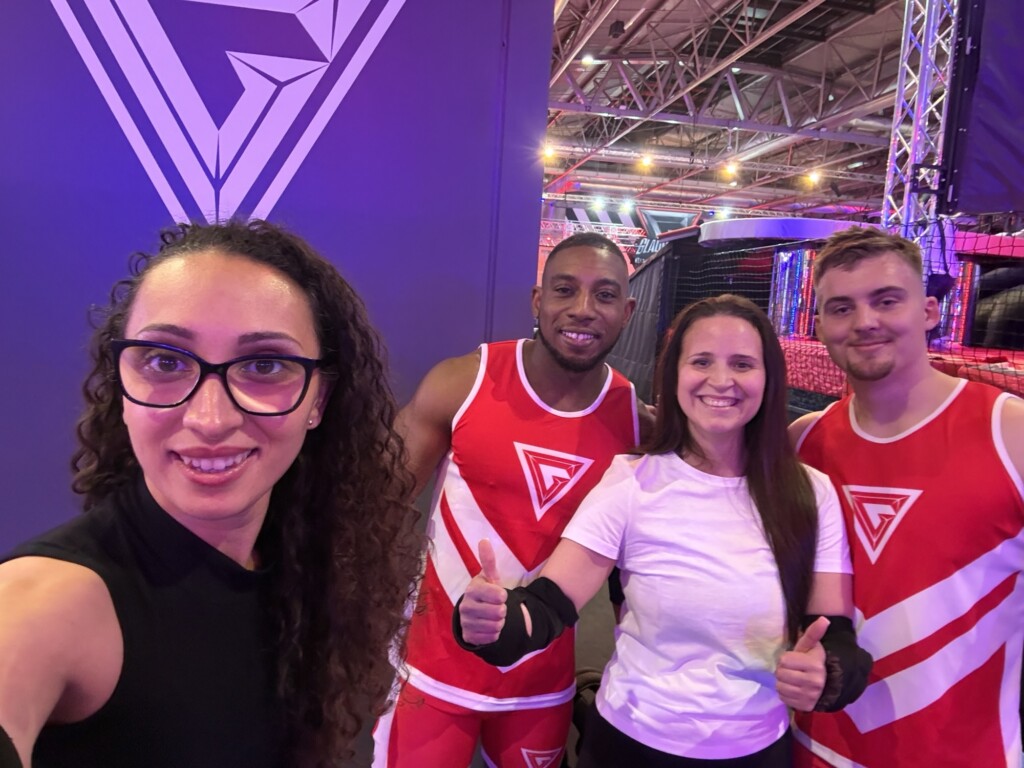 Four people smile for a group photo indoors; three wear red uniforms, one a white shirt, and two give a thumbs-up.