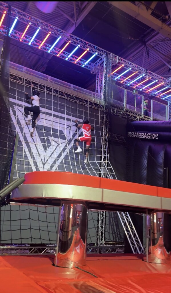 Two people are climbing a cargo net wall at an indoor obstacle course, with colourful lights shining overhead.