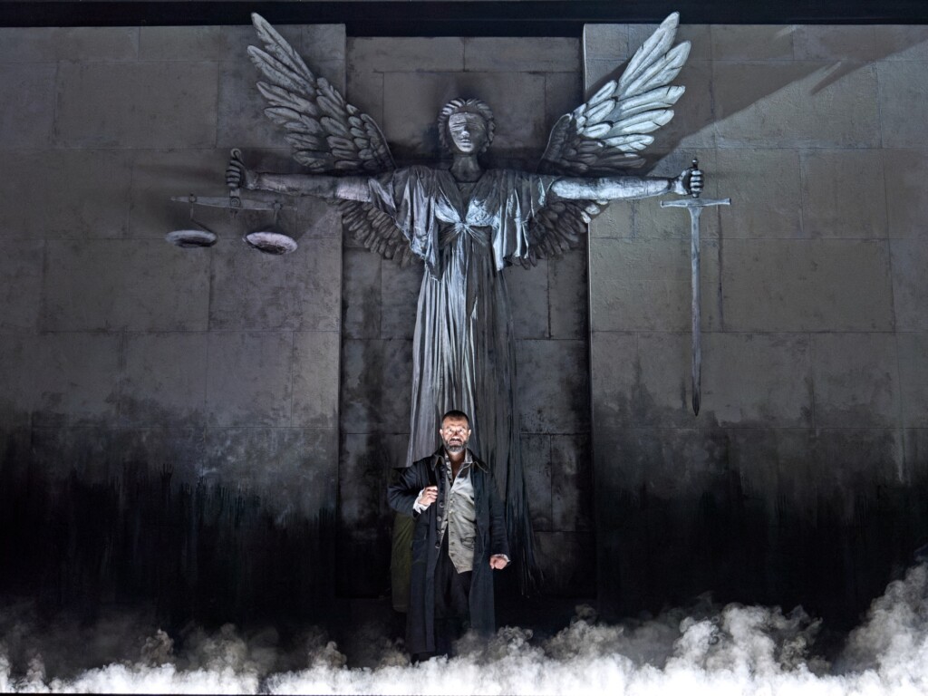 A man stands before a large statue of a winged figure holding scales and a sword, with mist at its base and a stone wall behind them.
