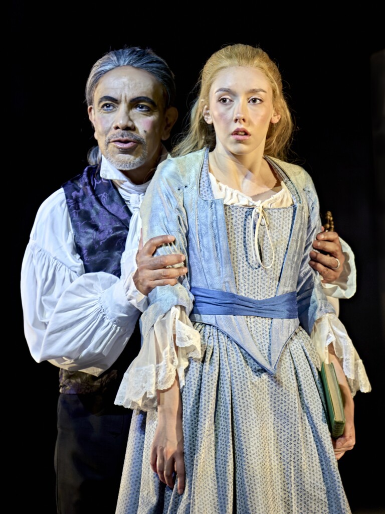 An older man in historical clothing holds a young woman in a blue dress, both staring ahead seriously against a dark backdrop.