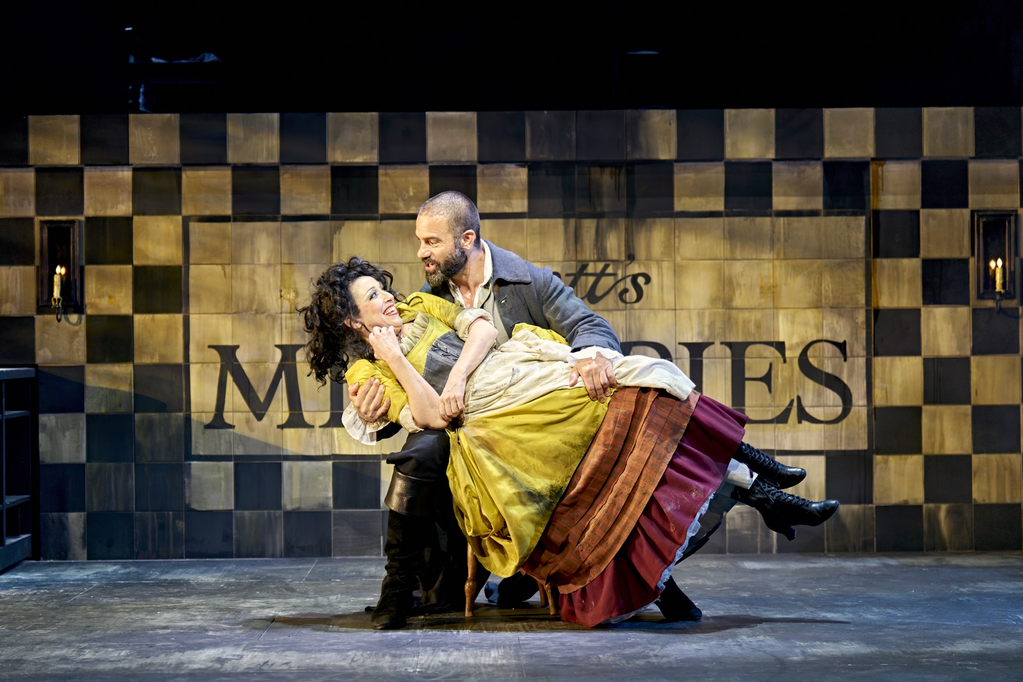 A man in old-fashioned clothing dips a smiling woman in a bright dress on stage, with a chequered wall and the word "MEMORIES" partly visible behind them.