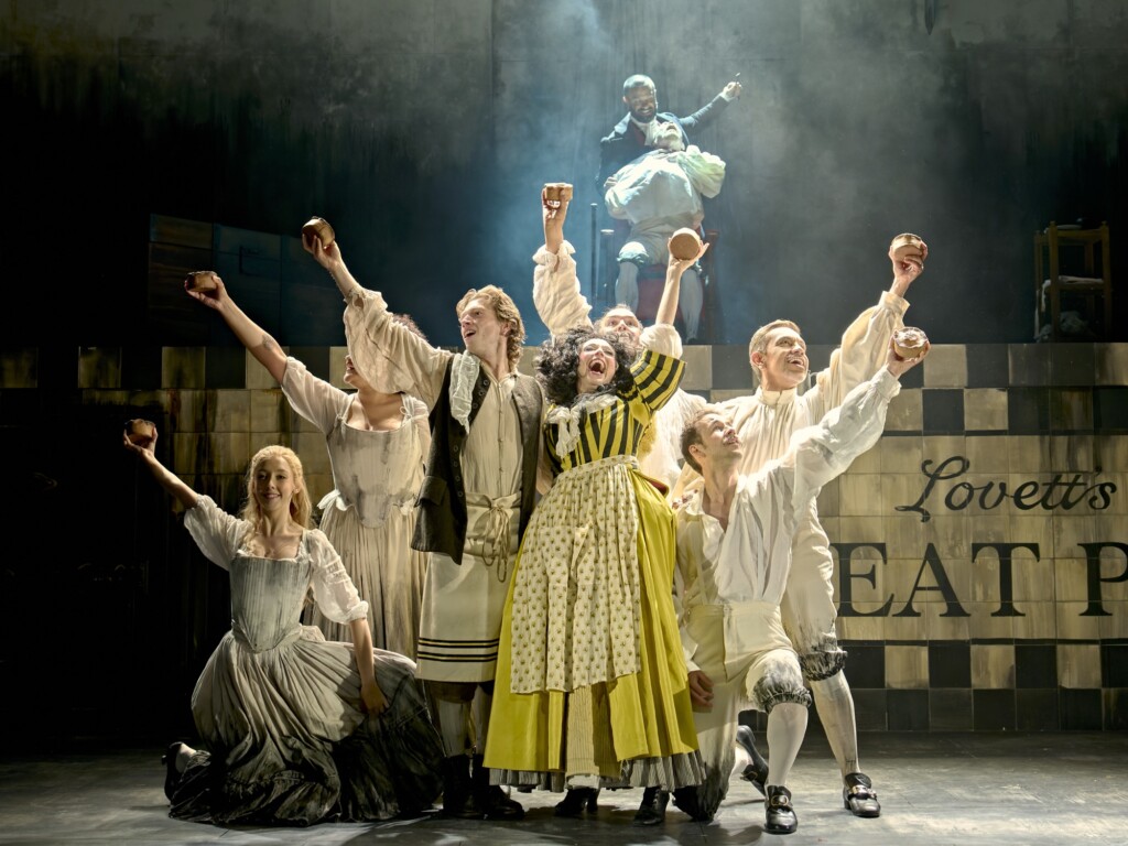 Actors in vintage costumes raise pies and tankards on stage, singing together in front of a sign for Lovett's Meat Pie bakery.