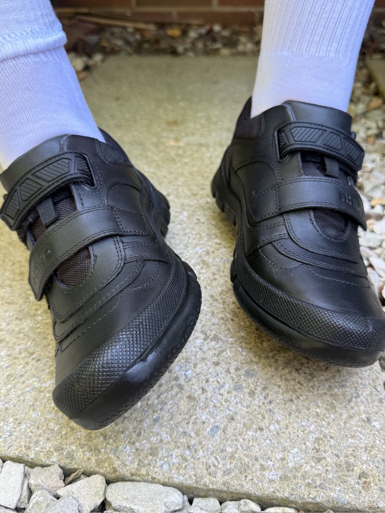 Someone in white socks and black velcro trainers stands on a concrete path lined with small stones.