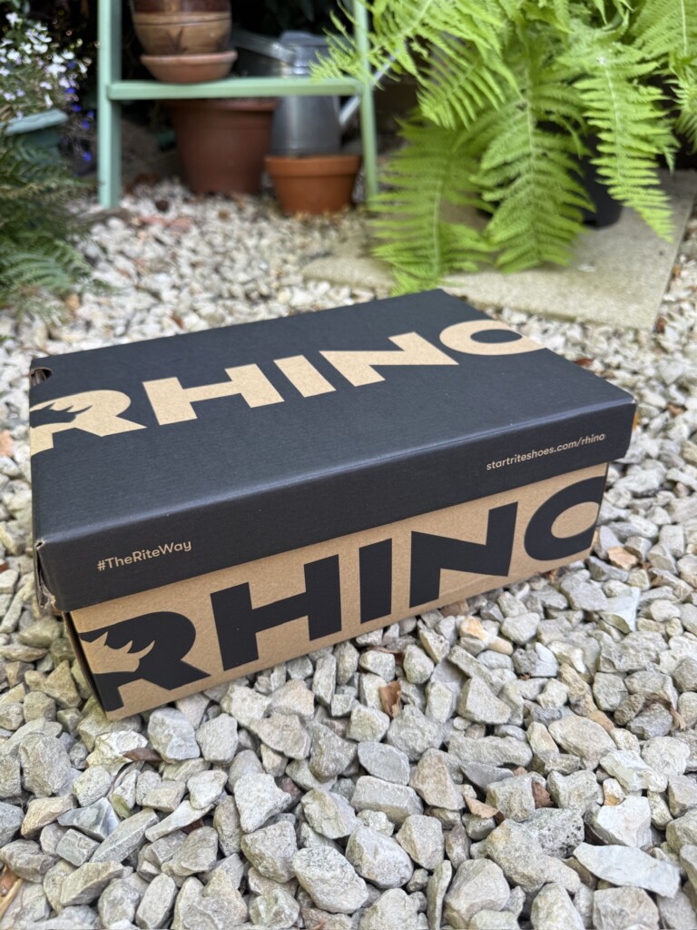 A cardboard box labelled “RHINO” rests on gravel outdoors, surrounded by green plants and potted ferns.