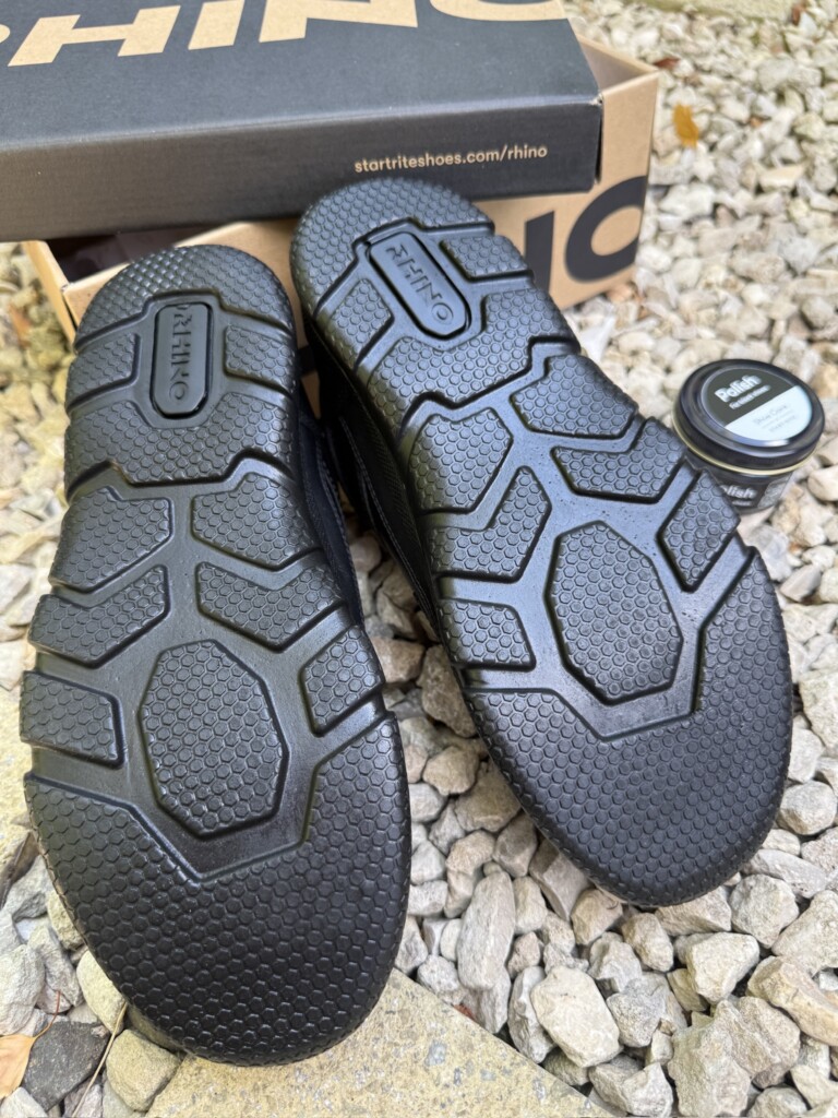 Two black school shoes lie sole-up on gravel next to a shoe polish tin and a “RHINO” shoe box.