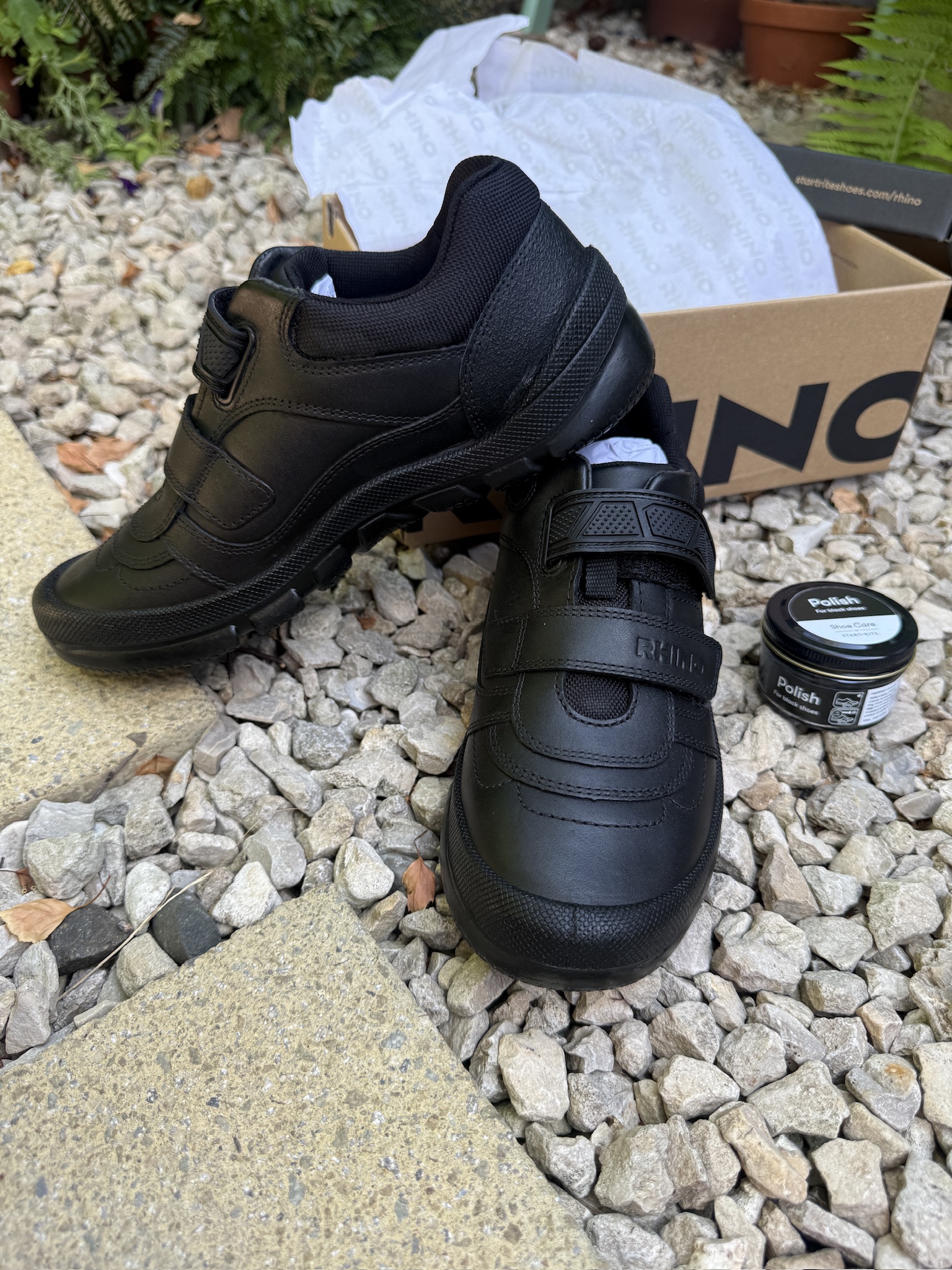 New black school shoes with Velcro straps are on gravel beside their box and a small tin of polish.