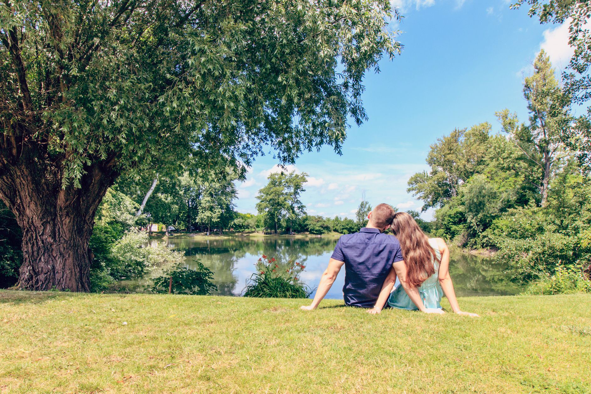 A couple sit on the grass beneath a tree, looking out at a pond.